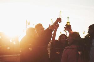 Beam of sunlight going into the camera. Holidays on the rooftop. Cheerful group of friends raised their hands up with alcohol photo