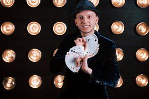 Your turn to pick. Magician in black suit and with playing cards standing in the room with special lighting at backstage