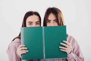 Hiding behind green notepad. Two sisters twins standing and posing in the studio with white background photo