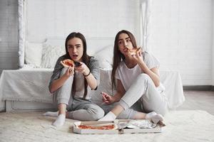 Food is always more delicious when watching movies. Sisters eating pizza in front of TV while sits on the floor of beautiful bedroom at daytime photo