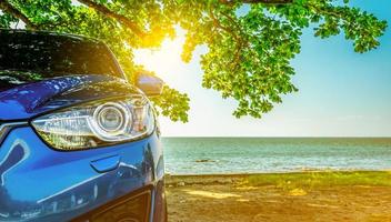 Blue sport SUV car parked by the tropical sea under umbrella tree. Summer vacation at the beach. Summer travel by car. Road trip. Automotive industry. Hybrid and electric car concept. Summer vibes. photo