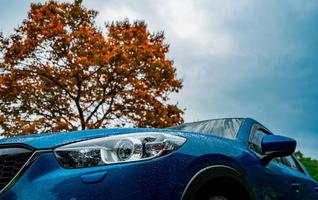 camión suv compacto azul con diseño deportivo y moderno estacionado cerca de un árbol caducifolio rojo con cielo azul y nubes. tecnología respetuosa con el medio ambiente. éxito empresarial y concepto de viaje. foto