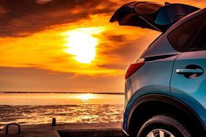 Blue compact SUV car with sport and modern design parked on concrete road by the sea at sunset. Environmentally friendly technology. Road trip travel on vacation at the beach and open car truck. photo