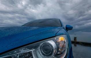 Selective focus on headlights of blue luxury SUV car parked on concrete road beside sea beach on rainy day with dark storm sky. Raindrops on blue SUV car with opened headlamp. Road trip travel. photo