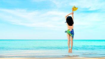 Happy young woman in pink swimwear hand holding hat and jumping at sand beach. Relaxing and enjoying holiday at tropical paradise sea beach. Girl in summer vacation. Summer vibes. Traveling alone. photo