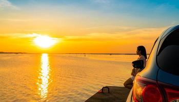 una joven asiática usa una camiseta, pantalones cortos y gafas de sol en la cabeza, apoyada en un camión deportivo azul compacto junto al mar al atardecer con hermosas nubes y un cielo amarillo anaranjado y reflejo en el agua foto