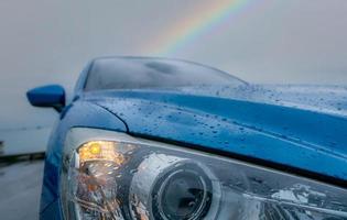 Front view of blue luxury SUV car parked near sea beach on rainy day with rainbow as background. Raindrops on blue texture of car bonnet. Car opened headlamp light in bad weather for safe driving. photo