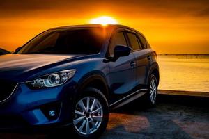 Front view blue compact SUV car with sport and modern design parked on concrete road by the sea at sunset. Electric car technology and business. Hybrid auto and automotive. Tropical road trip travel. photo