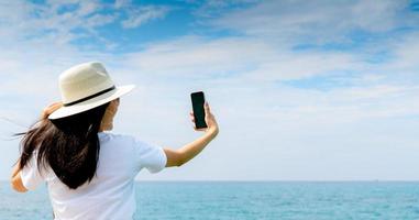 una joven asiática usa un sombrero en un estilo informal, usa un teléfono inteligente para tomar selfie en el muelle. vacaciones de verano en la playa del paraíso tropical. chica feliz viaje de vacaciones. la mujer disfruta y relaja la vida. vibras de verano. foto