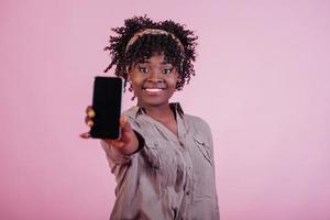 Holding black phone in hand. Attractive afro american woman in casual clothes at pink background in the studio photo