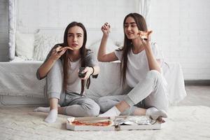 Girl searching something interesting. Sisters eating pizza when watching TV while sits on the floor of beautiful bedroom at daytime photo