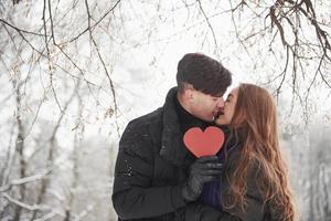 sosteniendo cartón rojo en forma de corazón. hermosa pareja joven se divierten juntos en el bosque nevado foto