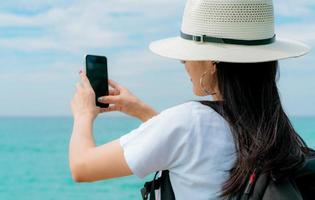 Young Asian backpacker woman wear hat use smartphone taking selfie at pier. Summer vacation at tropical paradise beach. Happy hipster girl travel on holiday. Woman enjoy and relax life. Summer vibes. photo