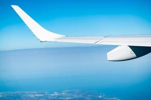 Wing of plane over the city and land. Airplane flying on blue sky. Scenic view from airplane window. Commercial airline flight. Plane wing above clouds. Flight mechanics concept. International flight. photo