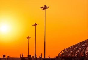 Tall electric pylon in the airport at sunset with an orange sky. Spotlights pole at the airfield. Energy support at the airport concept. Metal structure architecture. Outdoor LED lamp lights poles. photo