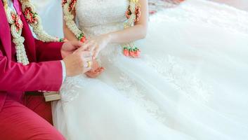 el novio puso el anillo de diamantes de la boda en el dedo de la novia para la propuesta el día de la ceremonia de la boda. novia y novio con vestido de novia blanco y traje rojo y guirnaldas de boda. casamiento o matrimonio. comenzar la vida de pareja. foto