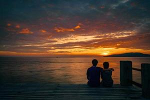 Silhouette of young romantic couple in love is sitting and hugging on wooden pier at the beach in sunrise time with golden sky. Vacation and travel concept. Romantic young couple dating at seaside.