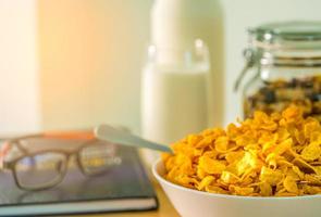 Bowl of cereal with spoon on the wood table on blurred background of glass of milk, bottle, glass container, textbook and eyeglasses. Calcium food for breakfast. Cornflakes and milk concept. photo