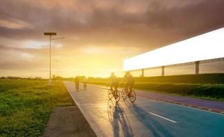 Sports man ride bicycles on the road in the evening near blank advertising billboard with sunset sky. Summer outdoor exercise for healthy and happy life. Cyclist riding mountain bike on bike lane.