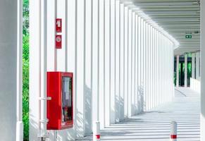 Empty walkway between buildings in the morning with shade of sunlight through space between white pole. Red fire extinguisher installed on concrete pole for safety. Modern architecture of corridor. photo