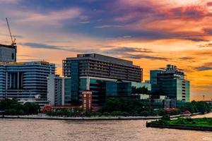 paisaje urbano de un edificio moderno cerca del río por la mañana al amanecer. edificio de oficinas de arquitectura moderna en tailandia. edificio frente al río y ciudad con cielo naranja y nubes foto
