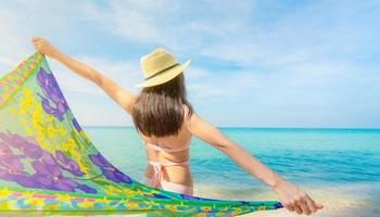 Back view of adult Asian woman wear pink bikini and straw hat relaxing and enjoying holiday at tropical paradise beach. Girl in summer vacation fashion. Summer vibes. Woman in swimwear arms outstretch