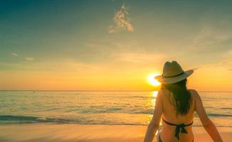 Back view of Asian woman wear bikini and straw hat sit on sand beach relax and enjoy holiday at tropical paradise beach. Woman watch beautiful sunset. Summer vacation. Travel alone. Summer vibes. photo