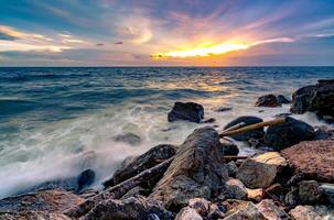 Ocean water splash on rock beach with beautiful sunset sky and clouds. Sea wave splashing on stone at sea shore on summer. Nature landscape. Tropical paradise beach at sunset. Rock beach at coast. photo