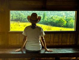 una joven asiática usa el sombrero sentada en un banco de madera y observa una hermosa vista del campo de hierba verde y el bosque en la torre de observación de la vida silvestre por la noche con la cálida luz del sol. concepto de viaje foto