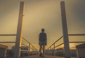 Asian Woman tourist walking alone with backpack on the bridge in the city in the evening on sunset with yellow sky. Walk away from the bad thing concept. Beautiful woman travel alone on her vacation photo