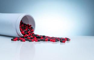 Red-black antibiotic capsule pills spread out of white plastic drug bottle on gradient background. Pharmaceutical industry. Antibiotic drug resistance concept. Healthcare and pharmaceutics concept. photo