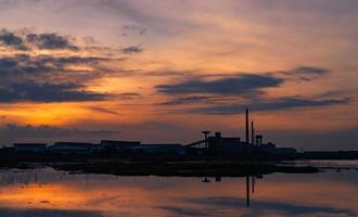 paisaje de edificios de la industria de la fábrica con reflejo azul oscuro y naranja del cielo de la puesta de sol sobre el agua en el río. edificio de almacén por la noche. ambiente limpio alrededor de la fábrica. fábrica cerrada. foto