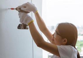 Asian woman drilling screw into apartment wall with cordless drill. Woman wear safety glasses and hand holding drilling tool for apartment renovation. Woman working home improvement with DIY concept.