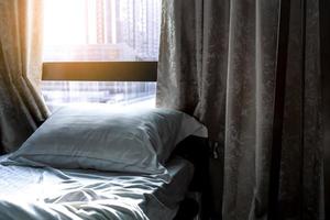 cama cómoda blanca y almohada suave en un dormitorio moderno. cama cerca de la ventana y cortina en el hotel por la mañana con luz solar. sábana de lino y funda de almohada. diseño de interiores de dormitorio. dormir con comodidad. foto