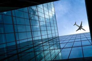 Airplane flying above modern glass office building. Perspective view of futuristic glass building. Exterior of office glass building. Business trip. Reflection in transparent glass. Company window. photo