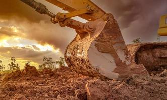 Dirt metal bucket of backhoe after digging soil. Backhoe parked at agricultural land on sunset sky background. Crawler excavator. Earthmoving machine at construction site at dusk. Excavation vehicle. photo