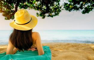 Back view of happy young Asian woman in pink swimsuit and straw hat relax and enjoy holiday at tropical sand beach under the tree. Girl in summer vacation fashion. Beauty sexy model. Summer vibes. photo