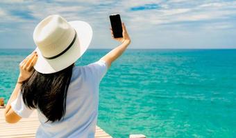 una joven asiática usa un sombrero de paja en un estilo informal, usa un teléfono inteligente para tomar selfie en un muelle de madera. vacaciones de verano en la playa del paraíso tropical. chica feliz viaje de vacaciones. la mujer disfruta y relaja la vida. foto