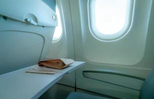 Plane window. Food in brown paper bag and plastic fork and spoon on plastic airplane tray table at seat back. Economy class airplane window. Inside of commercial airline. Seat with armchair. photo