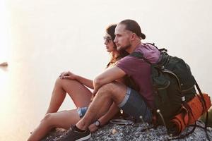 Hugging couple with backpack sitting on top of rock mountain enjoying view coast a river or lake. Traveling along mountains and coast, freedom and active lifestyle concept photo