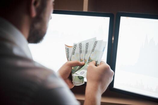 Man counts money in the office with multiple computer screens in index charts photo