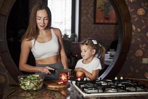 Having fun during the process. Pretty young woman standing in the modern kitchen near gas stove and teach daughter how to prepare food photo