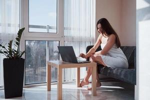 ciudad a través de la ventana. una mujer muy joven sentada en el sofá a la hora de la mañana en su habitación con una laptop foto