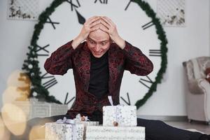 Too many presents. Man with big clock behind him sitting with New year gift boxes in holiday clothings photo