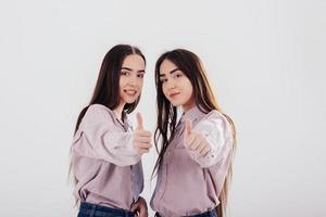 Girls approve by showing thumbs up. Two sisters twins standing and posing in the studio with white background photo