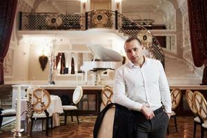 Portrait of guy in official wear stands in beautiful restaurant in vintage style with piano on the stage photo