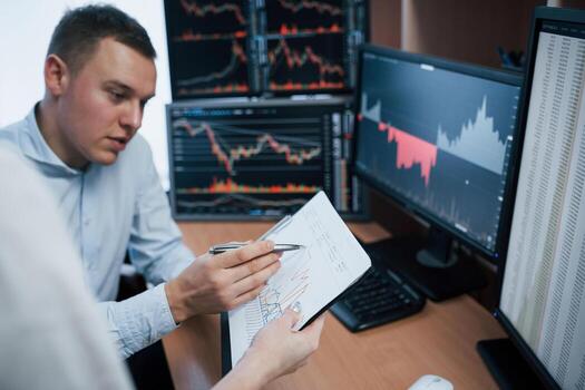 Notepad in hands with some graphs. Team of stockbrokers are having a conversation in a office with multiple display screens photo