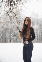 silencio de invierno. linda chica con cabello largo en blusa negra y anteojos está en el bosque nevado foto