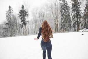 Rear view of girl with long hair runs near the forest to the automobile in wintertime photo