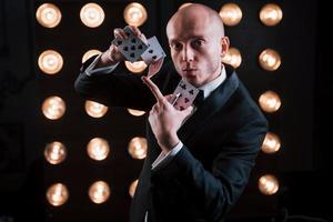 Loves his job of illusionist. Magician in black suit and with playing cards standing in the room with special lighting at backstage photo